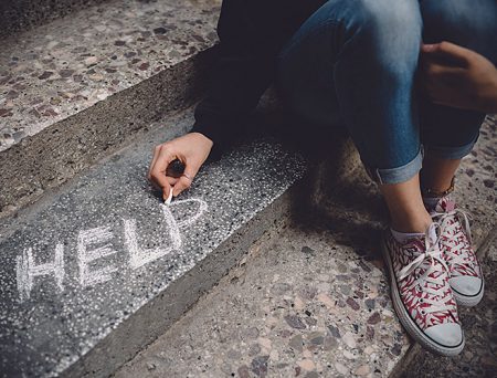 A young child is sitting on some concrete steps, writing "HELP" on one of them in white chalk.