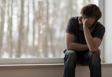 A young man in a dark gray shirt and blue jeans supports his head with his arm as he sits on a windowsill and stairs out of the window over his right shoulder.