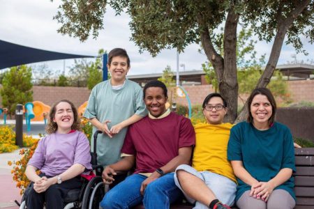 A diverse group of five adults sitting together outdoors. A diverse group of five adults sitting together outdoors on a bench and in a wheelchair, smiling at the camera under a tree with playground equipment visible in the background.