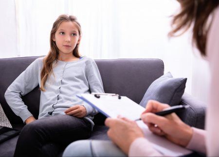 A young, teenage girl is sitting on a gray sofa with her legs crossed. She is looking at her therapist, who is blurred in the foreground, holding her clipboard and a pen.