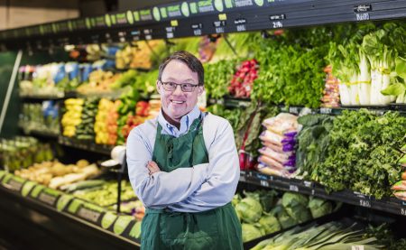 Smiling grocery store worker wearing glasses and a green apron. Smiling grocery store worker wearing glasses and a green apron stands with arms crossed in front of a colorful produce display of lettuce, peppers, and other vegetables.