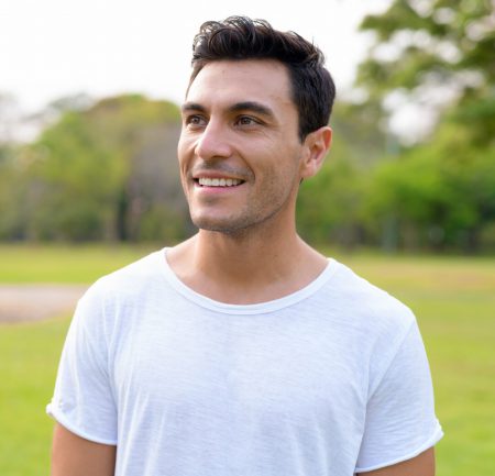 Picture of a young, handsome Hispanic man smiling. He is wearing a white t-shirt and appears to be at a public park in the summertime.