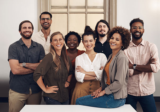 A group of eight young adults standing together and smiling at the camera in a bright indoor space. They appear to be a diverse team, wearing casual business and casual clothing, with some standing behind a low white table and others at the front. The setting suggests a professional but relaxed workplace environment.